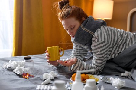 Photo for Redhead woman with cold blowing runny nose and swollen throat going to eat medicine, sitting on bed at home. lady in bedroom, with medicaments pills. ill female in domestic interior - Royalty Free Image