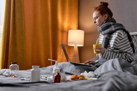 Foto de Mujer caucásica enferma tiene mala tos, sentado en la cama en casa, beber té caliente y trabajar en el ordenador portátil, trabajo en línea. Mujer joven con fuerte dolor de cabeza. covid-19, coronavirus, concepto omicrón - Imagen libre de derechos