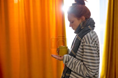 Photo for Young sick ginger redhead woman is treated and warmed by hot drink at home. Woman in scarf holding cup and pills, medicine in hands. Home treatment concept, close up side view portrait - Royalty Free Image