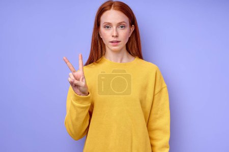 Foto de Hermosa mujer pelirroja seria en suéter elegante amarillo demostrando letra V símbolo de lenguaje de signos para sordos humanos con fondo azul. retrato de primer plano aislado - Imagen libre de derechos