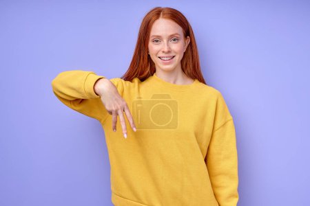 Foto de Hermosa mujer pelirroja alegre alegre en suéter elegante amarillo demostrando letra W símbolo de lenguaje de signos para sordos humanos con fondo azul. retrato de primer plano aislado - Imagen libre de derechos