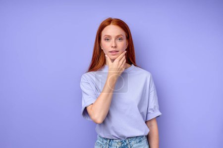 Photo for Beautiful ginger girl with hand on her chin looking at camera, red-haired girl has idea, plan, touching her chin, close up portrait isolated blue background - Royalty Free Image
