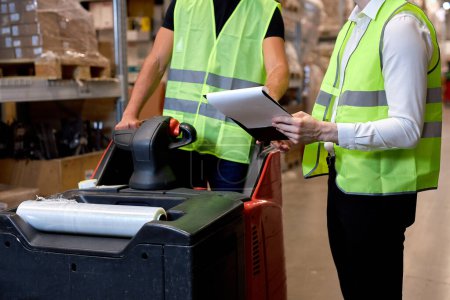 Photo for Warehouse workers men in reflective jackets pulling a pallet truck and talking while standing among shelves in retail warehouse logistics, distribution center. male holding clipboard, cargo control - Royalty Free Image