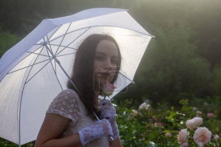 Foto de Una mujer elegante y clásica con un paraguas blanco sostiene una rosa bajo la luz del sol. Lleva un vestido de encaje vintage, rodeado de rosas en verano.. - Imagen libre de derechos