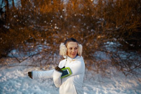 Téléchargez les photos : Jeune femme souriante veste blanche avec oreille plus chaude étirant à l'extérieur dans le parc de la ville en hiver. - en image libre de droit