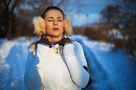 Téléchargez les photos : Femme en bonne santé en veste blanche avec oreille chaud reprendre haleine après l'exercice à l'extérieur dans le parc de la ville en hiver. - en image libre de droit