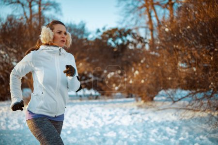 Téléchargez les photos : Femme active en veste blanche avec oreille plus chaude jogging à l'extérieur dans le parc de la ville en hiver. - en image libre de droit