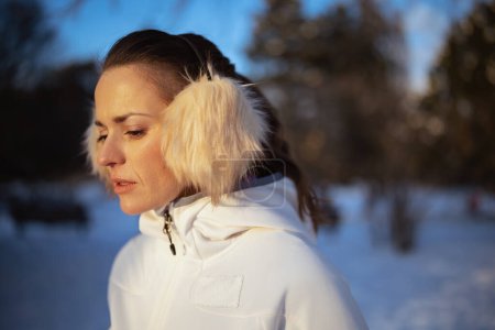 Téléchargez les photos : Femme active en veste blanche avec oreille plus chaude à l'extérieur dans le parc de la ville en hiver. - en image libre de droit