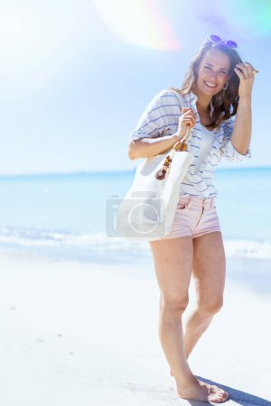 Photo for Full length portrait of smiling elegant woman with white straw bag in white striped shirt and shorts at the beach. - Royalty Free Image