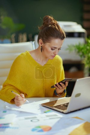 Foto de Pensativo moderno propietario de la pequeña empresa mujer en suéter amarillo con el ordenador portátil utilizando el teléfono inteligente en la oficina verde moderna. - Imagen libre de derechos