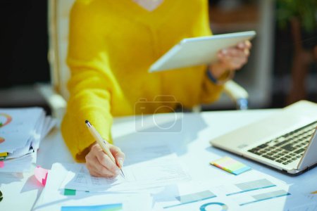 Foto de Primer plano de la mujer propietaria de una pequeña empresa en suéter amarillo usando tableta PC y trabajando con documentos en la oficina verde. - Imagen libre de derechos