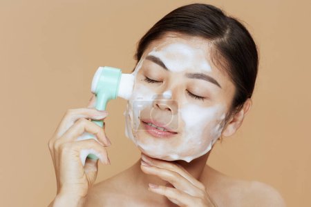 Foto de Relajada mujer moderna con masajeador lavando la cara contra fondo beige. - Imagen libre de derechos