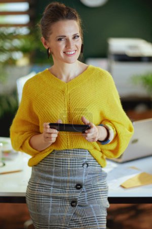 Téléchargez les photos : Heureuse femme propriétaire de petite entreprise d'âge moyen à la mode en pull jaune avec smartphone dans le bureau vert moderne. - en image libre de droit