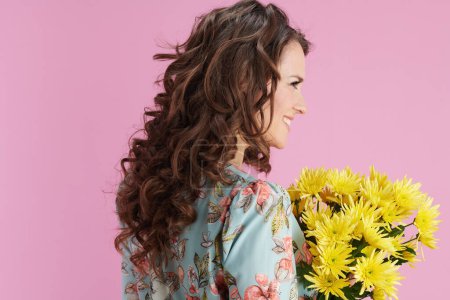 Téléchargez les photos : Souriant élégant femme d'âge moyen avec de longs cheveux bruns ondulés avec des fleurs de chrysanthèmes jaunes sur fond rose. - en image libre de droit
