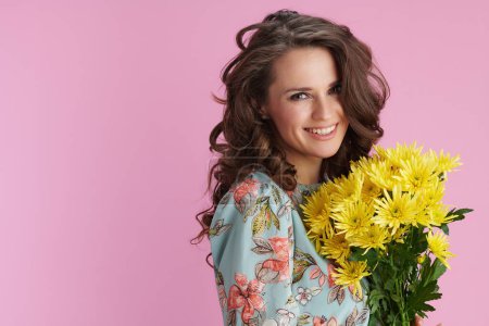 Foto de Retrato de mujer moderna sonriente en vestido floral con flores de crisantemos amarillos aisladas sobre fondo rosa. - Imagen libre de derechos