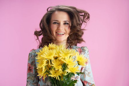 Photo for Smiling trendy woman with long wavy brunette hair with yellow chrysanthemums flowers jumping against pink background. - Royalty Free Image