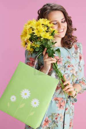 Foto de Feliz elegante mujer de 40 años en vestido floral con flores de crisantemos amarillos y bolsa verde aislada sobre fondo rosa. - Imagen libre de derechos