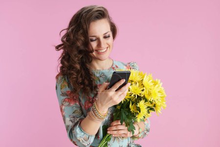 Photo for Happy young woman with long wavy brunette hair with yellow chrysanthemums flowers using smartphone app isolated on pink. - Royalty Free Image
