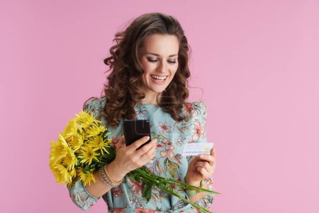 Téléchargez les photos : Heureuse élégante femme d'âge moyen en robe florale avec des fleurs de chrysanthèmes jaunes et carte de crédit en utilisant des applications smartphone isolé sur rose. - en image libre de droit