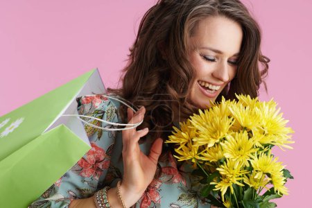 Photo for Happy stylish female in floral dress with yellow chrysanthemums flowers and green shopping bag isolated on pink background. - Royalty Free Image