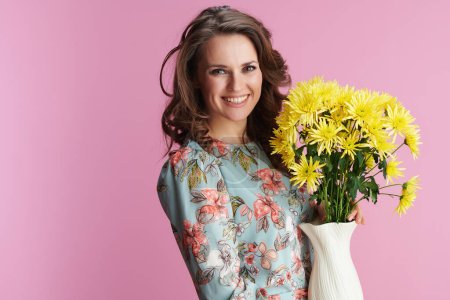 Téléchargez les photos : Portrait de heureuse femme moderne en robe florale avec des fleurs de chrysanthèmes jaunes dans un vase isolé sur rose. - en image libre de droit