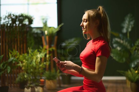 Foto de Mujer moderna en ropa de fitness roja en casa verde moderna haciendo yoga y usando teléfono inteligente. - Imagen libre de derechos