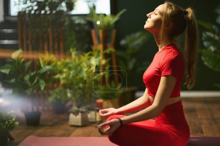 Foto de Mujer joven en ropa de fitness roja en el moderno invernadero meditando. - Imagen libre de derechos