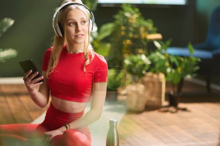Photo for Young woman in red fitness clothes using smartphone and listening to the music with headphones at modern green home. - Royalty Free Image