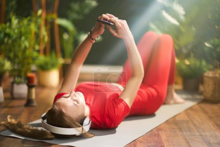 Téléchargez les photos : Jeune femme en rouge vêtements de fitness avec tapis de yoga à l'aide d'un smartphone et l'écoute de la musique avec écouteurs à la maison verte moderne. - en image libre de droit