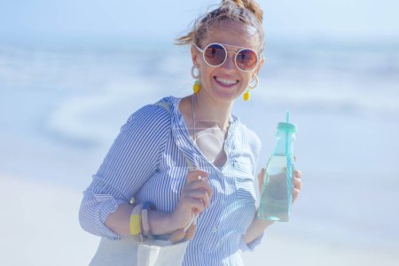 Téléchargez les photos : Portrait de femme souriante moderne de 40 ans avec sac en paille blanche et boisson à la plage. - en image libre de droit