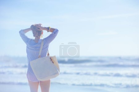 Foto de Visto desde atrás femenino con bolsa de paja blanca relajándose en la playa. - Imagen libre de derechos