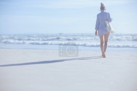 Téléchargez les photos : Vu de derrière femelle avec sac de paille blanche marchant sur la plage. - en image libre de droit