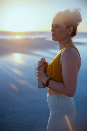 Foto de Mujer deportiva en ropa deportiva con botella de agua y rastreador de fitness en la playa al atardecer. - Imagen libre de derechos