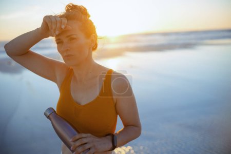 Téléchargez les photos : Fatigué femme sportive saine en vêtements de sport avec bouteille d'eau à la plage le soir. - en image libre de droit