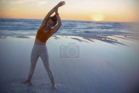 Foto de Retrato de larga duración de la mujer deportiva sana en ropa de fitness en la playa por la noche estiramiento. - Imagen libre de derechos