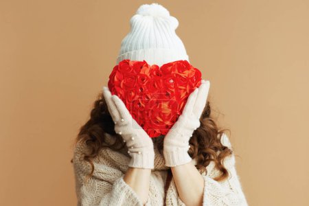 Téléchargez les photos : Bonjour l'hiver. femme tendance en pull beige, mitaines et chapeau sur fond beige avec coeur rouge. - en image libre de droit