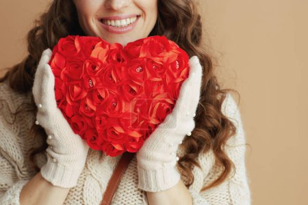 Photo for Hello winter. Closeup on happy woman in beige sweater, mittens and hat isolated on beige background with red heart. - Royalty Free Image
