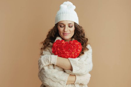 Téléchargez les photos : Bonjour l'hiver. femme élégante détendue en pull beige, mitaines et chapeau sur fond beige avec coeur rouge. - en image libre de droit