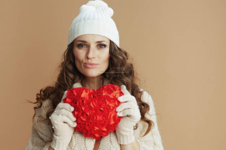 Foto de Hola invierno. Retrato de mujer elegante en suéter beige, mitones y sombrero con corazón rojo aislado en beige. - Imagen libre de derechos