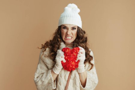 Photo for Hello winter. angry stylish woman in beige sweater, mittens and hat isolated on beige background with red heart. - Royalty Free Image