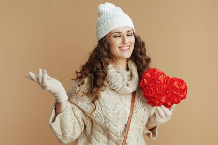 Photo for Hello winter. happy stylish woman in beige sweater, mittens and hat against beige background with red heart. - Royalty Free Image