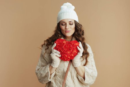 Photo for Hello winter. smiling stylish middle aged woman in beige sweater, mittens and hat isolated on beige background with red heart. - Royalty Free Image