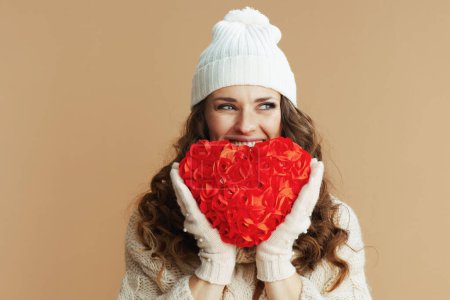 Foto de Hola invierno. feliz mujer moderna de 40 años en suéter beige, mitones y sombrero aislados sobre fondo beige con corazón rojo. - Imagen libre de derechos
