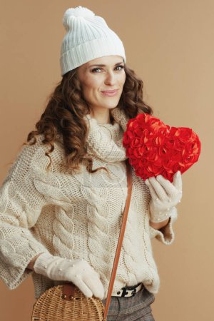 Photo for Hello winter. happy stylish middle aged woman in beige sweater, mittens and hat isolated on beige background with red heart and handbag. - Royalty Free Image