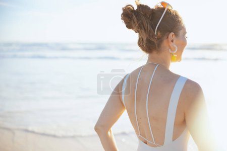Foto de Visto desde atrás elegante mujer de mediana edad en ropa de playa blanca en la playa relajante. - Imagen libre de derechos