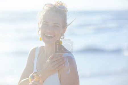 Photo for Portrait of happy elegant woman in white swimwear at the beach applying sunscreen. - Royalty Free Image