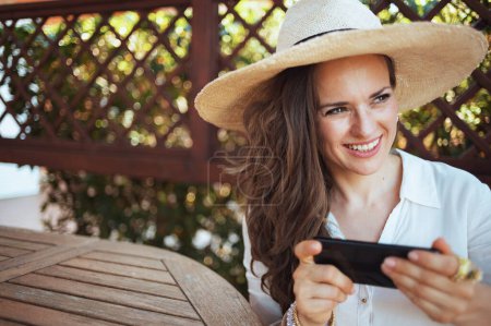 Téléchargez les photos : Heureuse jeune femme en chemise blanche avec chapeau assis à la table envoyer un message texte en utilisant smartphone sur la terrasse de l'hôtel maison d'hôtes. - en image libre de droit
