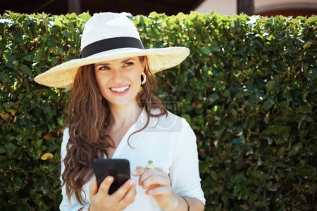 Foto de Ama de casa elegante feliz en camisa blanca con sombrero usando aplicaciones de teléfonos inteligentes al aire libre cerca de la pared verde. - Imagen libre de derechos