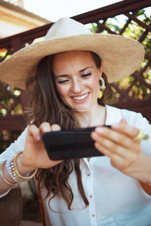 Téléchargez les photos : Souriant élégant femme au foyer de 40 ans en chemise blanche avec chapeau envoyer un message texte en utilisant smartphone sur la terrasse de l'hôtel maison d'hôtes. - en image libre de droit
