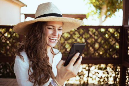 Téléchargez les photos : Souriant tendance 40 ans femme au foyer en chemise blanche avec chapeau ayant vidéo réunion sur un smartphone sur la terrasse de l'hôtel maison d'hôtes. - en image libre de droit